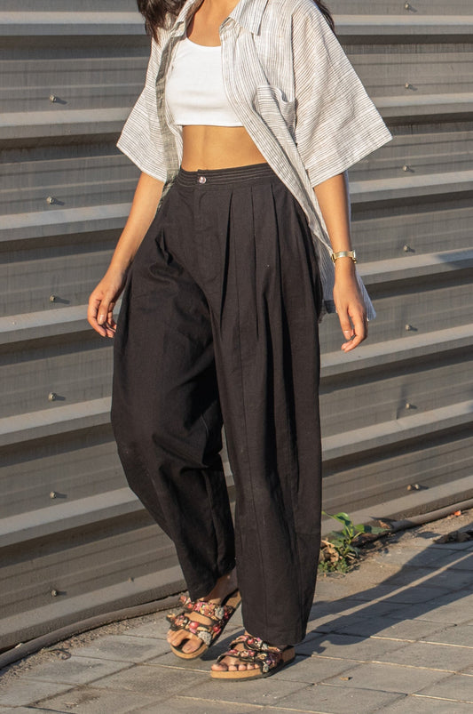 Woman wearing a white striped shirt, white top, and black barrel leg pleated pants standing against a corrugated metal wall.