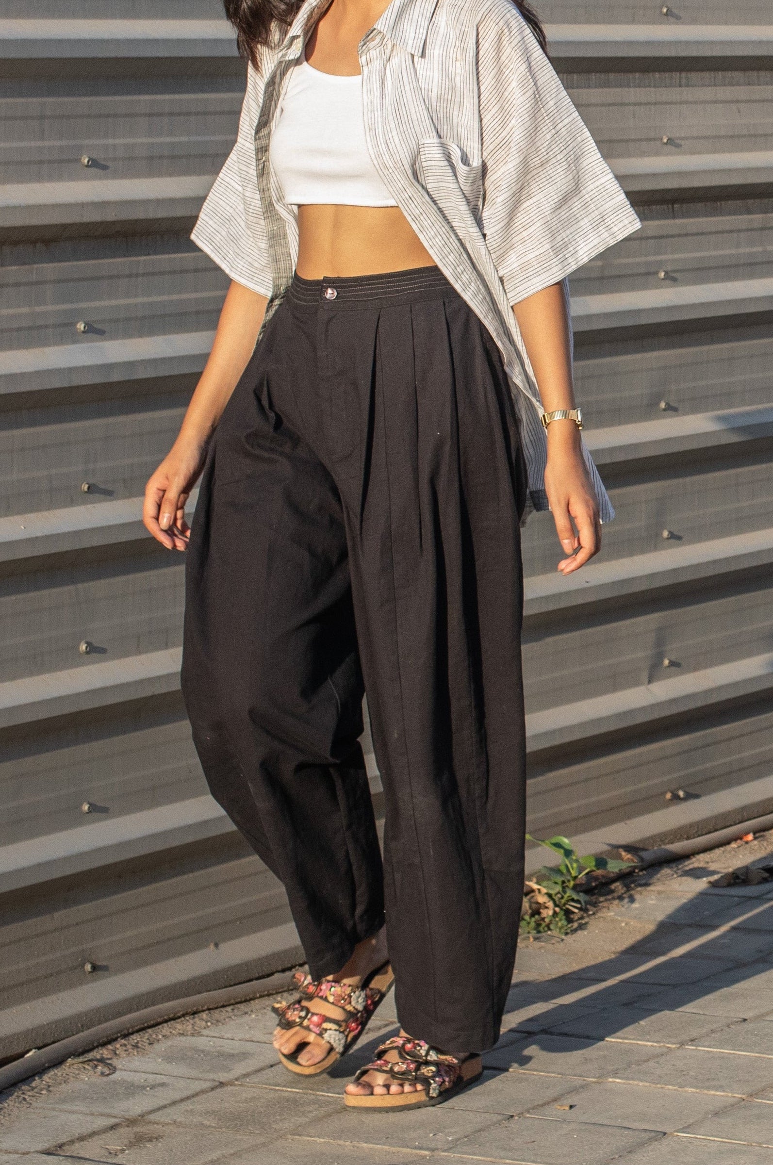 Woman wearing a white striped shirt, white top, and black barrel leg pleated pants standing against a corrugated metal wall.